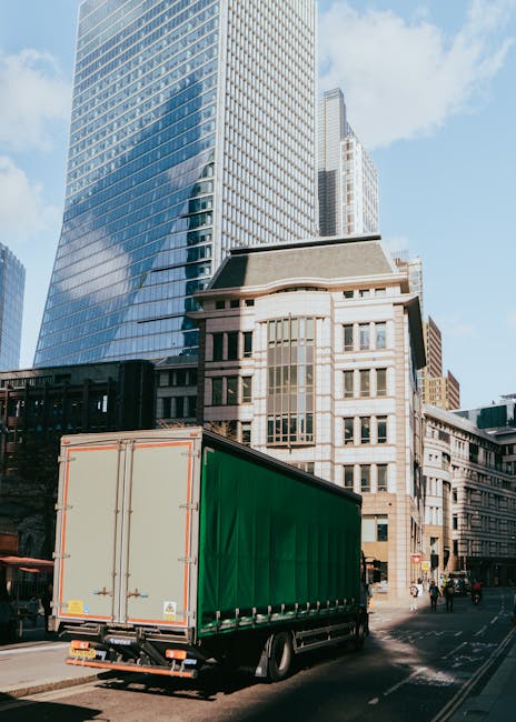 A narrow street scene showing a white moving truck parked between two multi-storey buildings with light-colored facades and wooden window shutters. The truck, positioned on the pavement, is being load