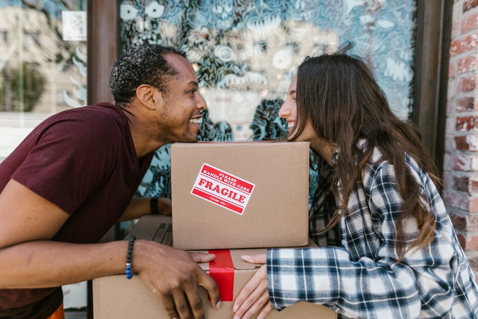 A man and a woman are outdoors during a house relocation process, each holding a cardboard box wrapped with red packing tape and marked with a red 'Fragile' label. The man, with short curly hair and wearing a maroon t-shirt, is leaning forward with a smile, engaging with the woman. The woman has long brown hair and is dressed in a plaid shirt, also smiling and facing the man closely. They appear to be exchanging boxes or preparing to lift them together. Behind them is a large window with decorative blue and white patterned film, suggesting they are near the entrance of a house, possibly on a pavement or driveway. Visible around them are other packing materials, possibly additional boxes or protective coverings. This image captures a moment of packing and moving, linking to the detailed home relocation and furniture transport services offered by Man and a Van Harringay, who specialise in removals in tight urban environments like Harringay Ladder.