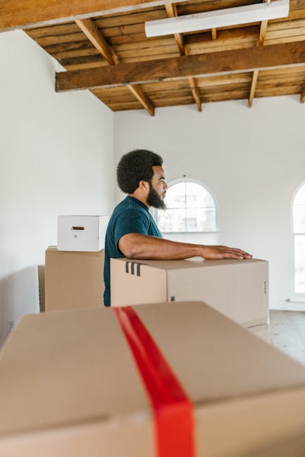 A man with dark curly hair and a beard, wearing a blue T-shirt, is standing inside a spacious room with white walls and large arched windows. He is positioned behind several cardboard boxes, some sealed with red packing tape, which are stacked on the floor. The room has an unfinished look with a wooden ceiling featuring exposed beams and minimal lighting. The boxes appear to be part of a home relocation process, possibly being prepared for transport or unpacked after moving. The man is inspecting or arranging the boxes as part of packing and moving activities. This scene reflects the logistics involved in a furniture transport or household removal service, as provided by Man and a Van Harringay, which specializes in house removals and packing assistance in Harringay.