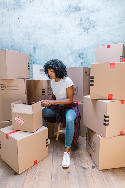 A man and a woman are outdoors during a house relocation process, each holding a cardboard box wrapped with red packing tape and marked with a red 'Fragile' label. The man, with short curly hair and wearing a maroon t-shirt, is leaning forward with a smile, engaging with the woman. The woman has long brown hair and is dressed in a plaid shirt, also smiling and facing the man closely. They appear to be exchanging boxes or preparing to lift them together. Behind them is a large window with decorative blue and white patterned film, suggesting they are near the entrance of a house, possibly on a pavement or driveway. Visible around them are other packing materials, possibly additional boxes or protective coverings. This image captures a moment of packing and moving, linking to the detailed home relocation and furniture transport services offered by Man and a Van Harringay, who specialise in removals in tight urban environments like Harringay Ladder.