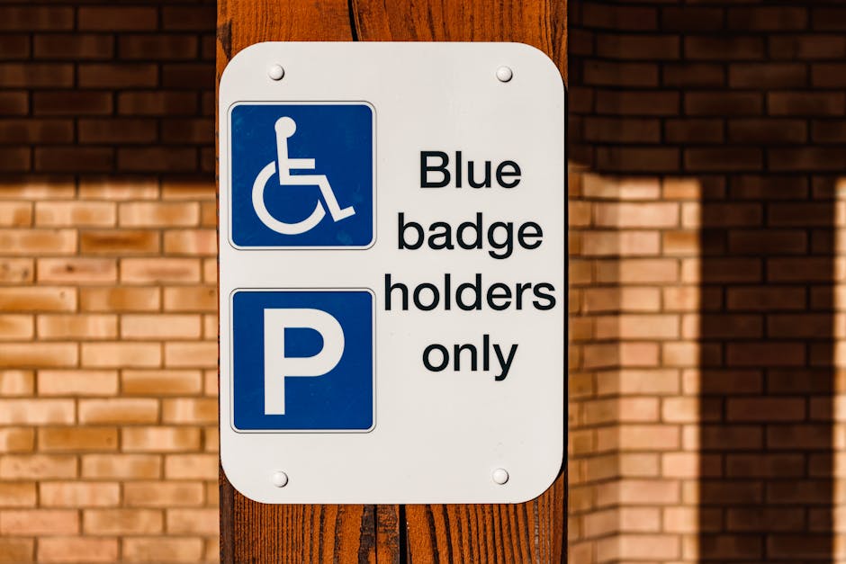 A rectangular white parking sign mounted on a wooden post in front of a brick building wall. The upper section of the sign displays a blue square with a white wheelchair icon indicating accessibility,